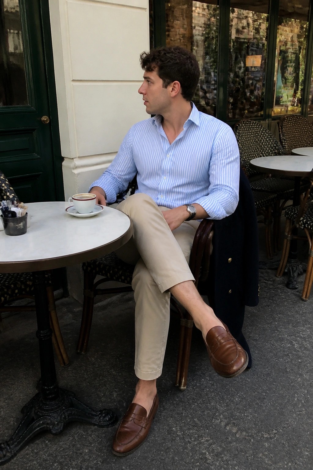 Man seated at cafe table wearing light blue striped oxford shirt, beige khaki pants, brown leather loafers, with navy coat draped nearby.