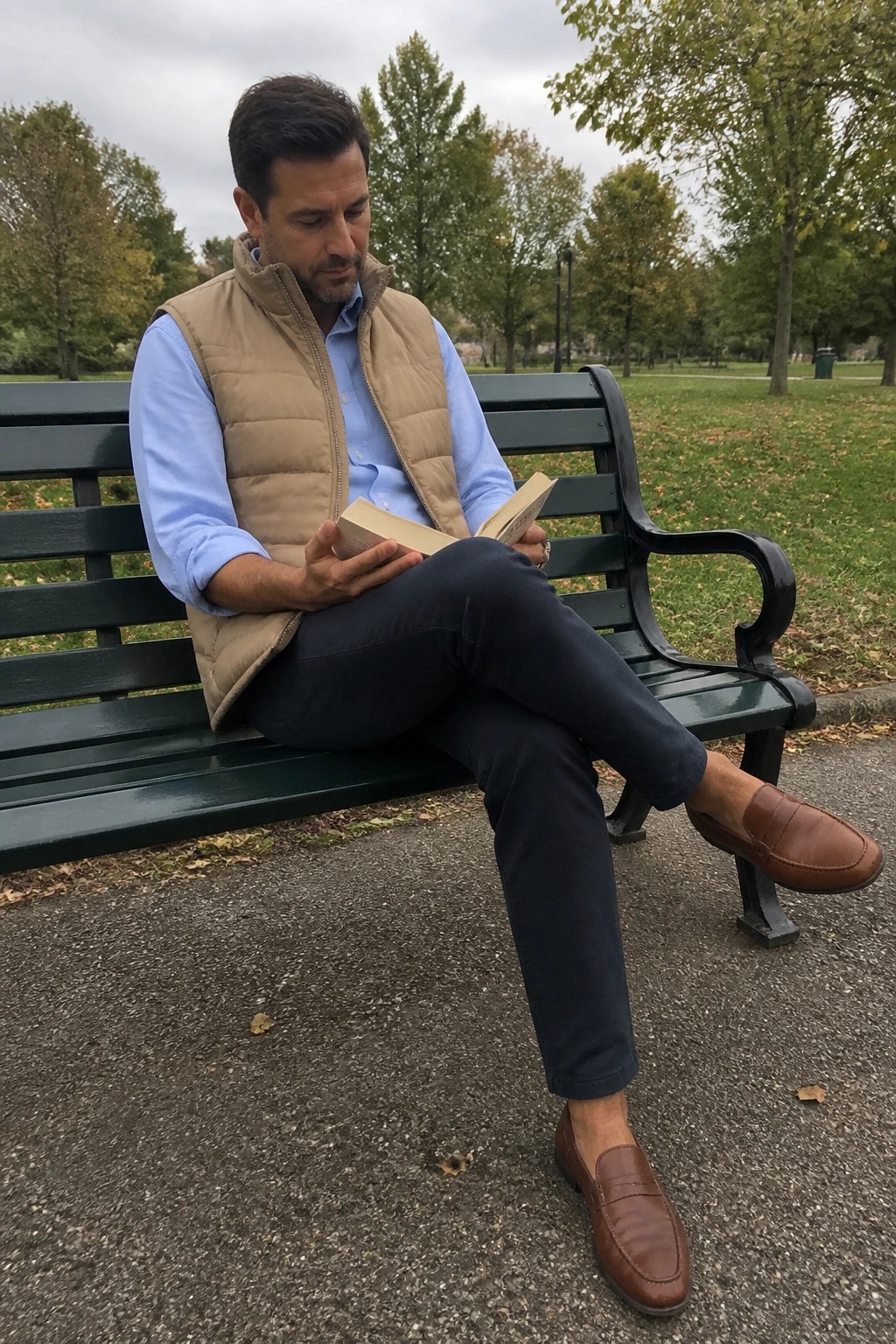 Man in beige vest and blue shirt reading book on green park bench