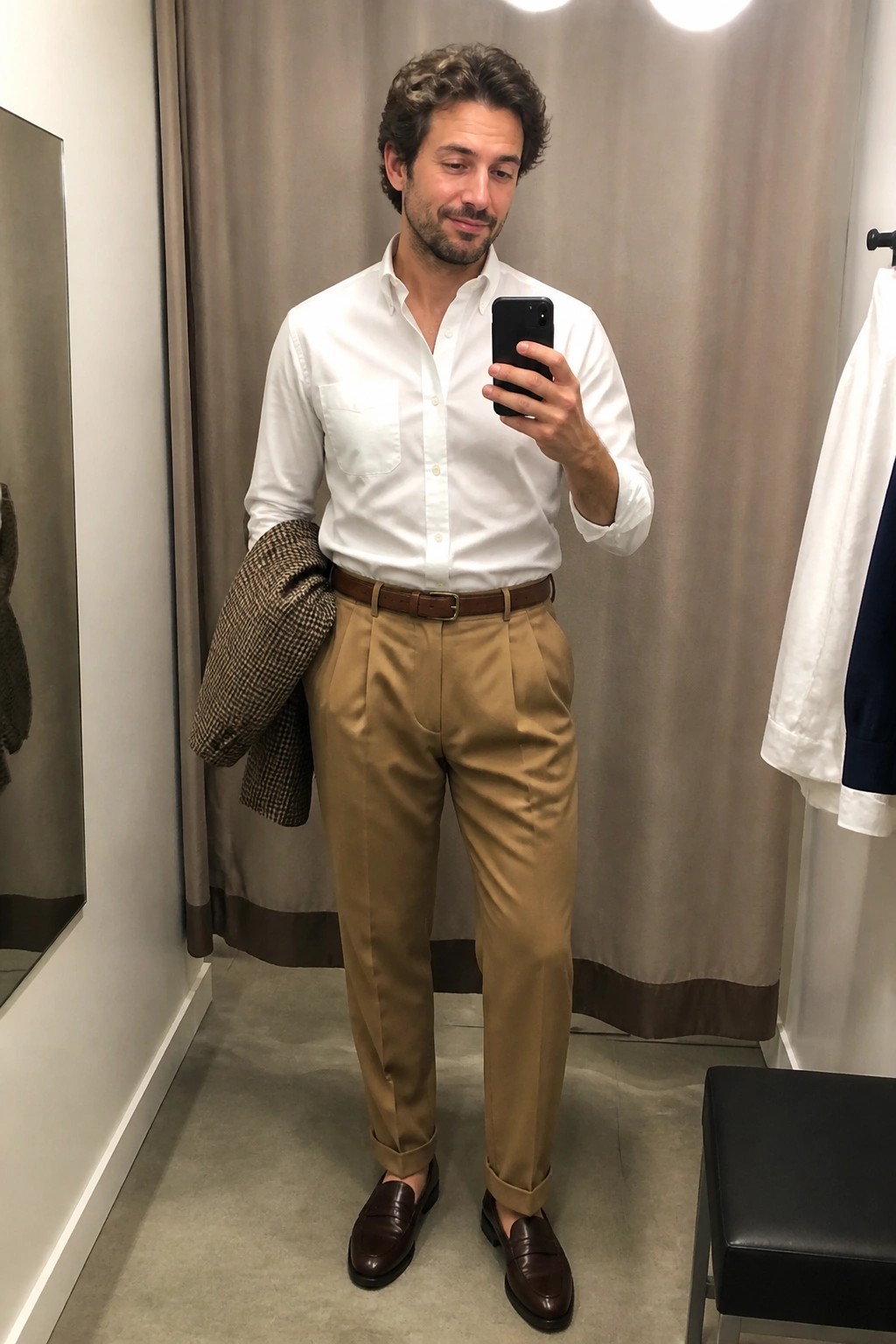 Man in white oxford shirt tucked into tan pleated trousers with brown belt and matching penny loafers holding a houndstooth jacket over one arm