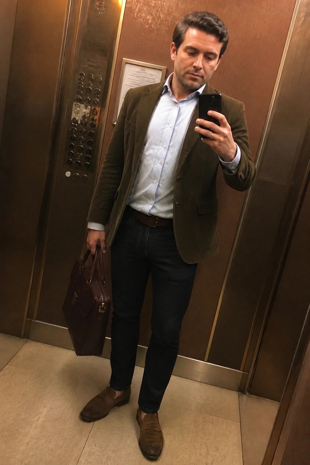 Man in green corduroy blazer taking mirror selfie in elevator holding briefcase