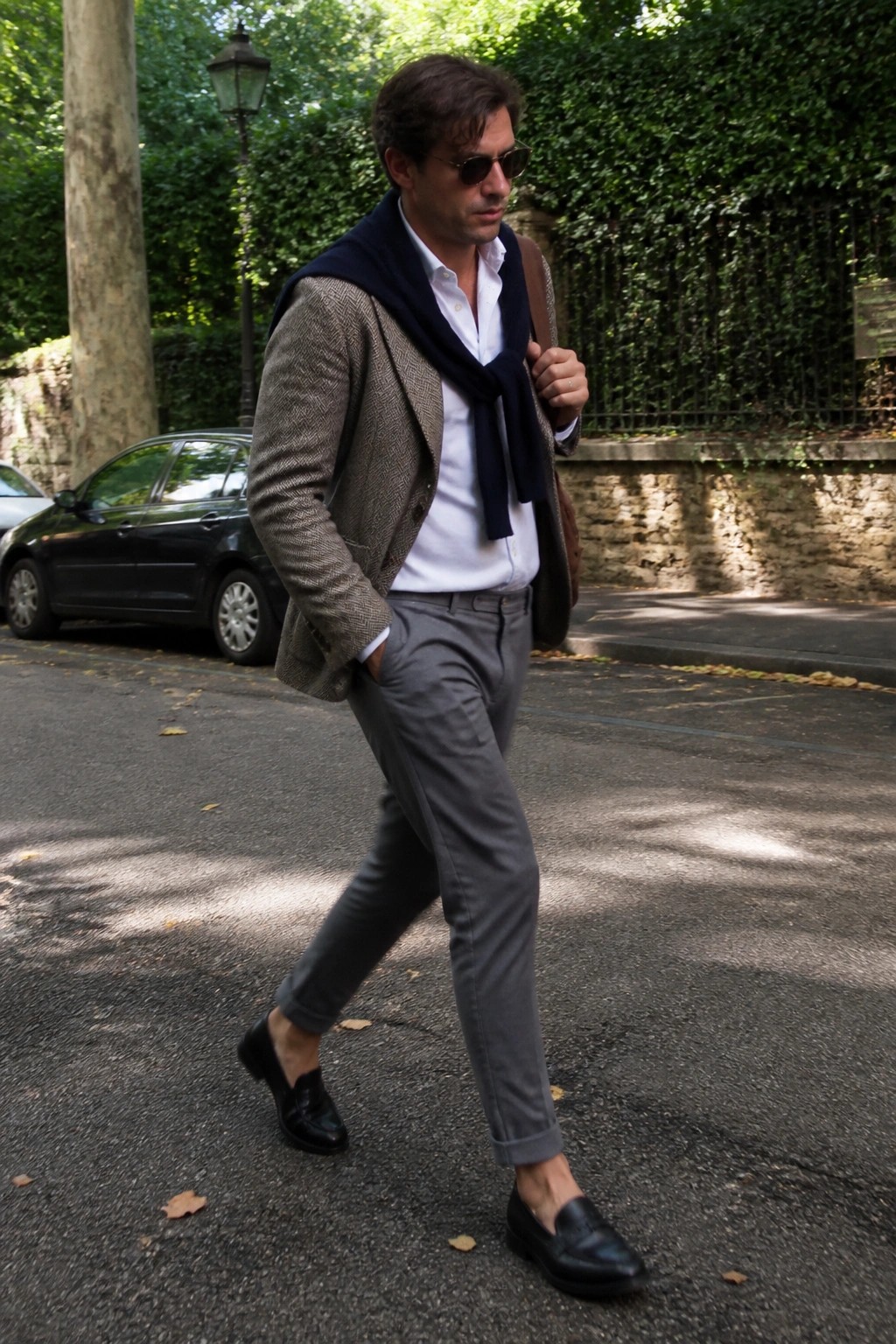 Man in herringbone blazer, white shirt, navy scarf, and gray trousers walking outdoors