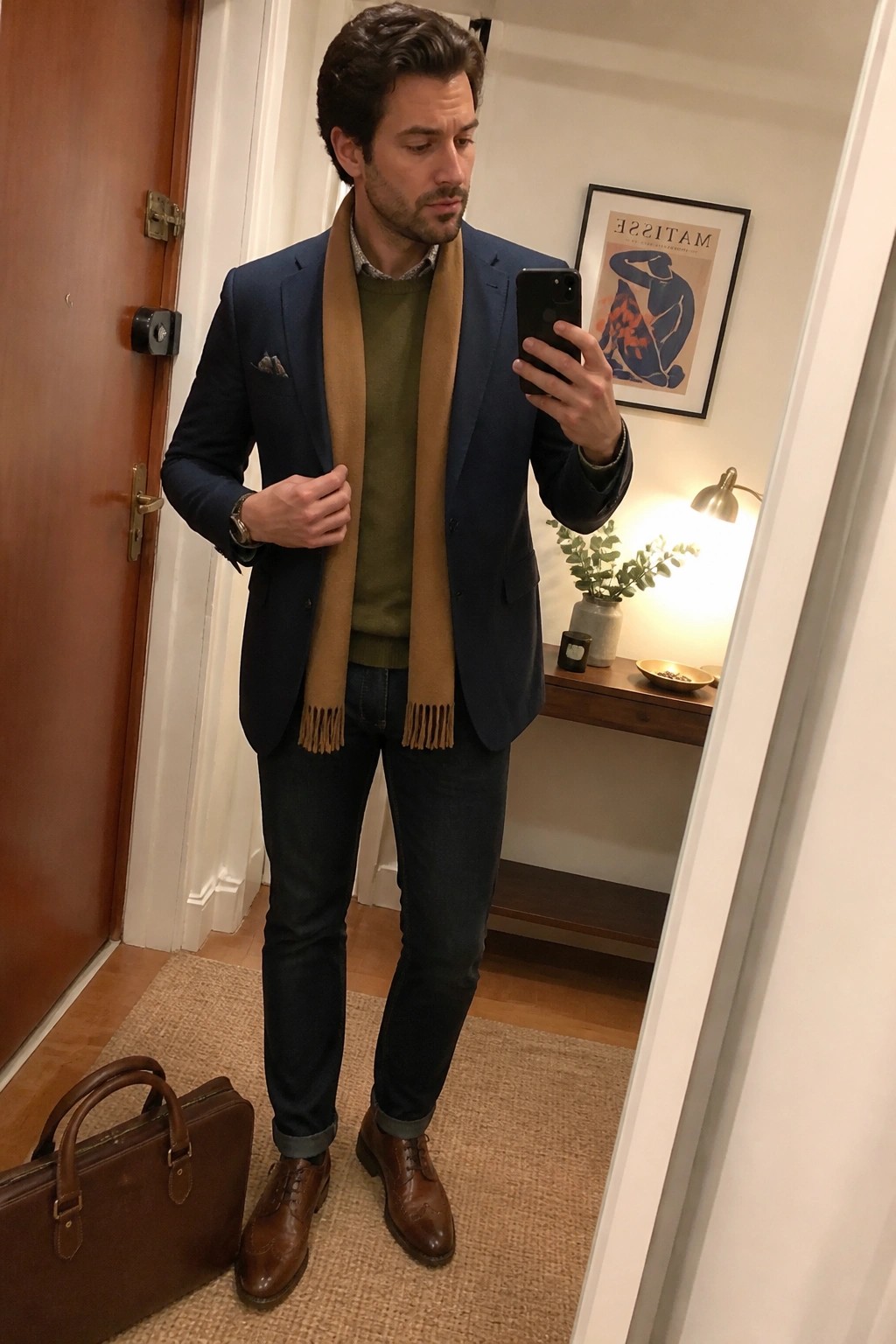 Man in navy blazer and tan scarf takes mirror selfie beside leather briefcase.