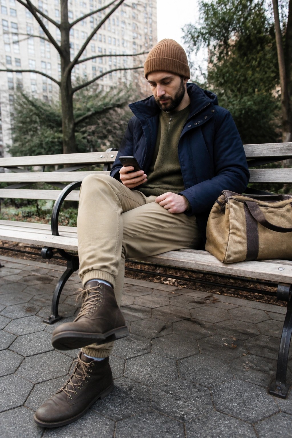 Bearded man in brown beanie and navy jacket views smartphone on park bench beside duffel bag.