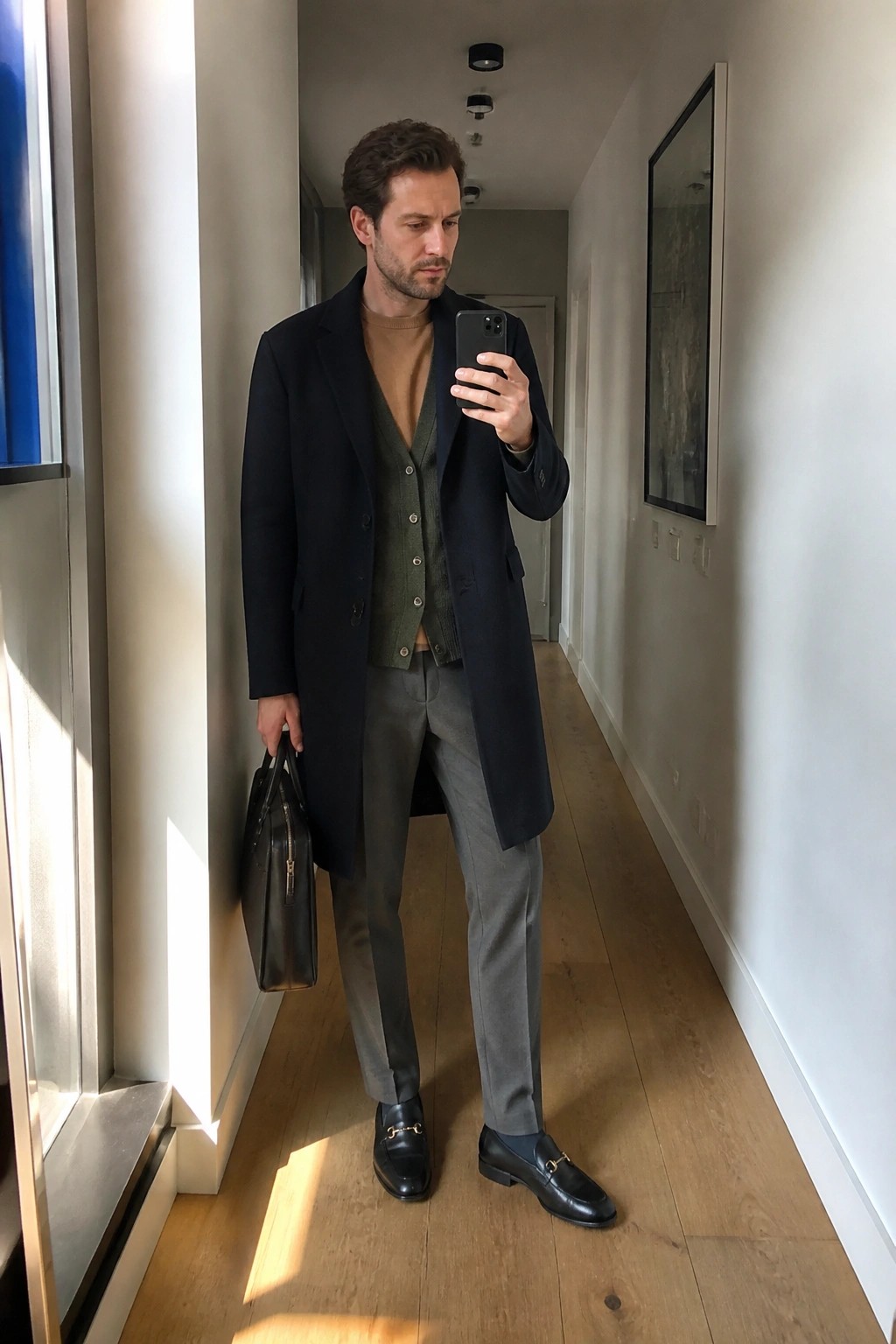 Man in navy overcoat and green sweater vest taking mirror selfie with briefcase