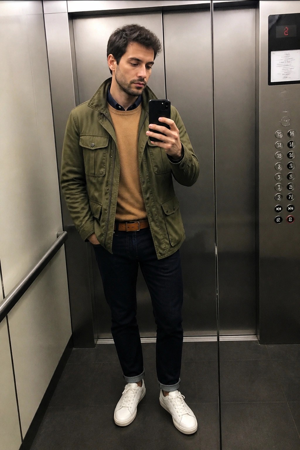 Man in green jacket over tan sweater taking selfie with phone in elevator