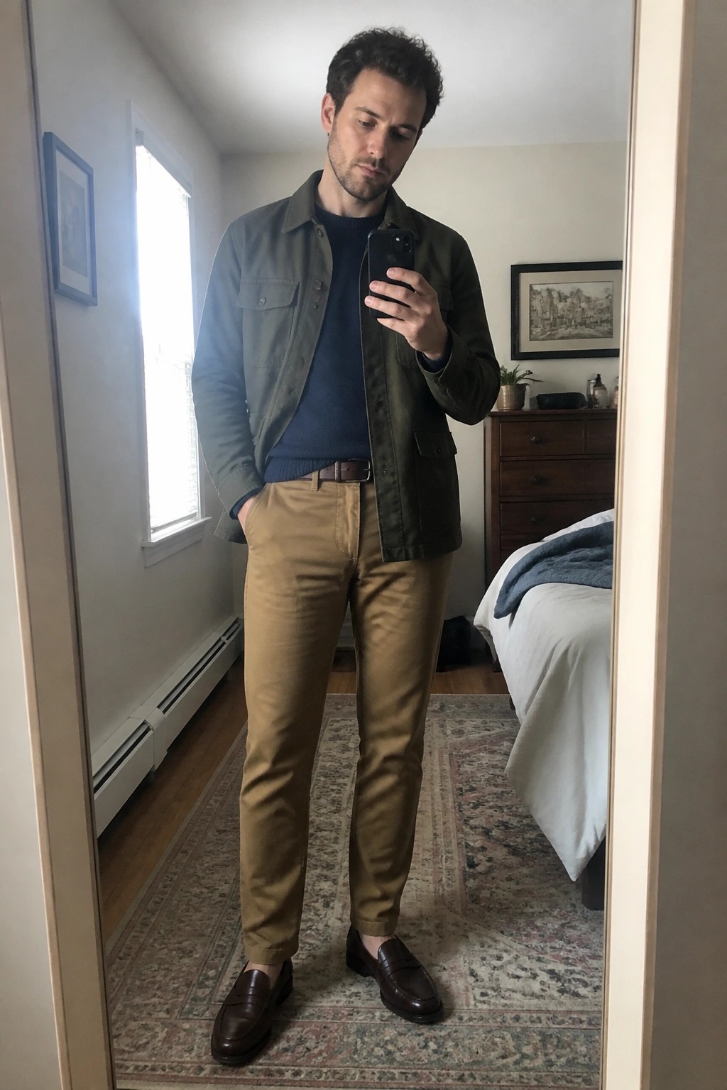 Man in green jacket and tan pants takes mirror selfie in bedroom.