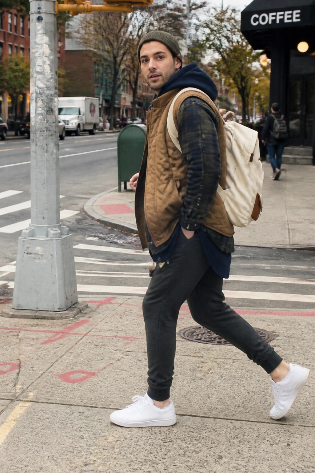 Man in green beanie, brown vest, and backpack crossing urban street crosswalk.