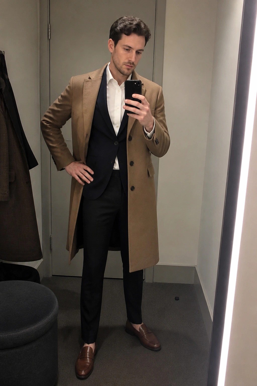 Man in tan overcoat over navy suit taking mirror selfie with phone