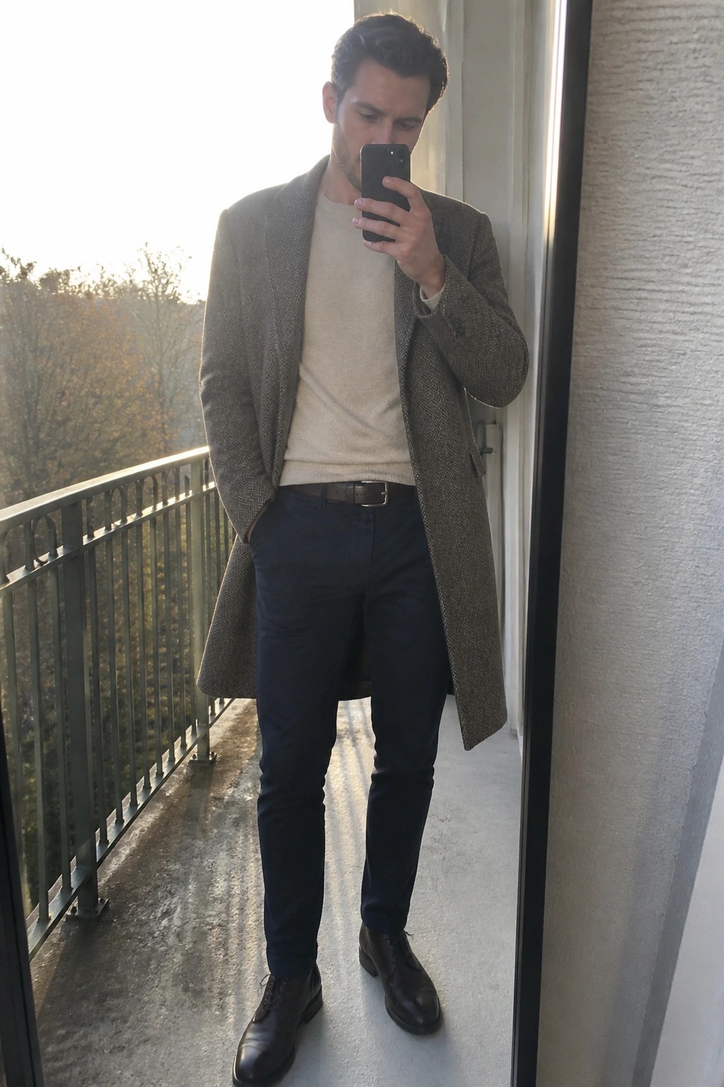 Man in grey herringbone coat over beige sweater taking balcony mirror selfie