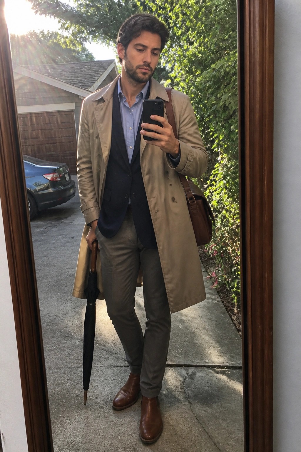 Man in beige trench coat and navy vest taking mirror selfie with phone and umbrella.