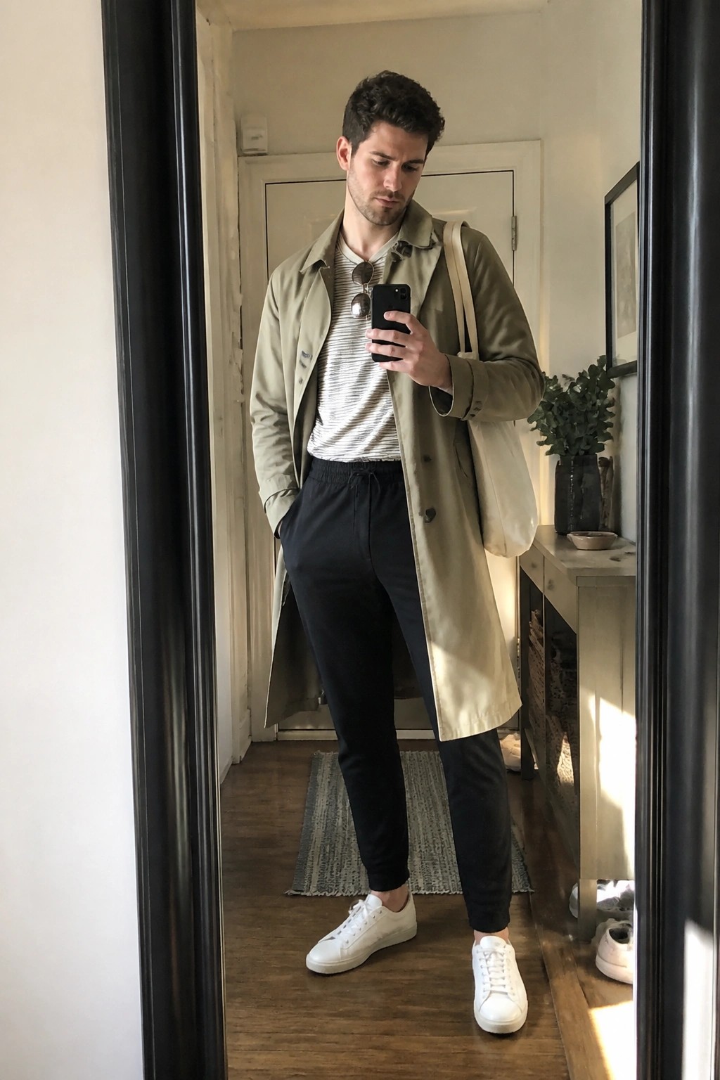 Man in beige trench coat and striped shirt takes mirror selfie with phone