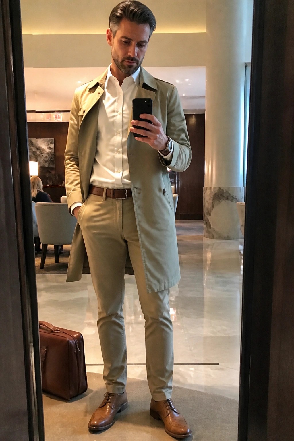Man in light trench coat and khaki pants taking mirror selfie in marble lobby