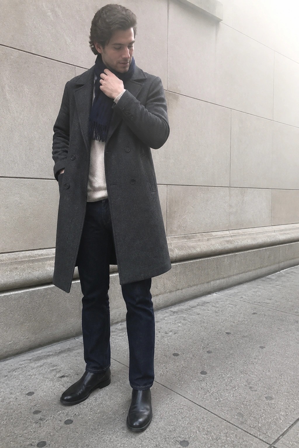 Tall man wearing a long dark gray double-breasted wool overcoat, navy fringed scarf, cream sweater, dark jeans, and black Chelsea boots standing against a stone wall.
