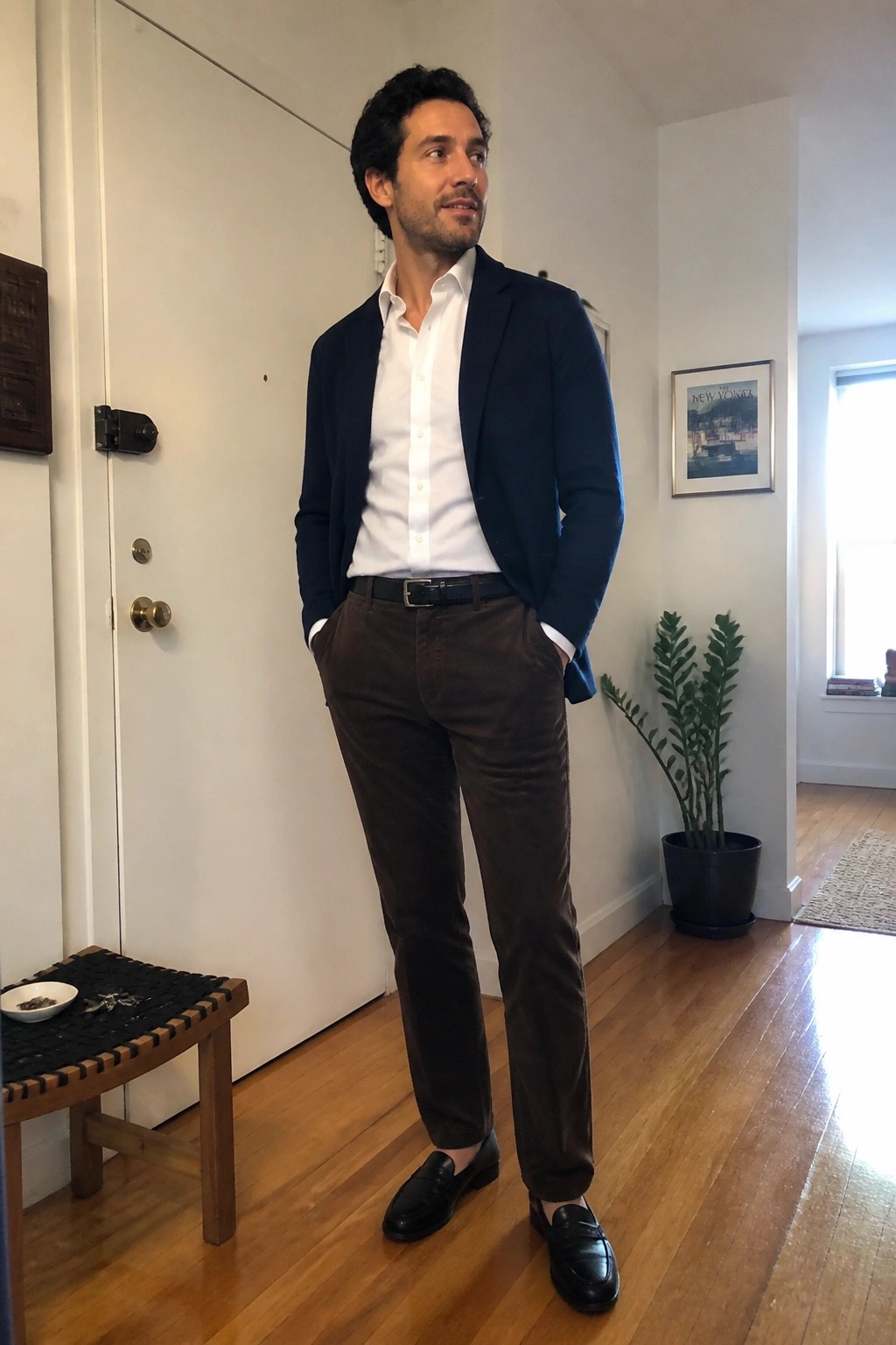 Man in navy blazer over white button down shirt with brown corduroy trousers and black loafers standing in a hallway.
