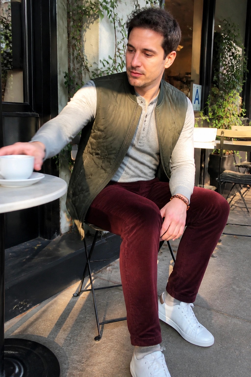 Man in green vest and maroon corduroy pants reaches for coffee at outdoor cafe.