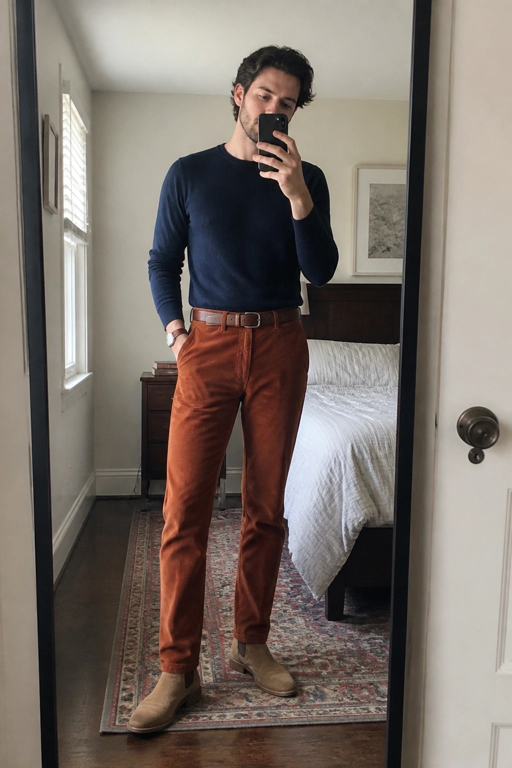 Man in navy sweater and rust corduroy pants taking bedroom mirror selfie