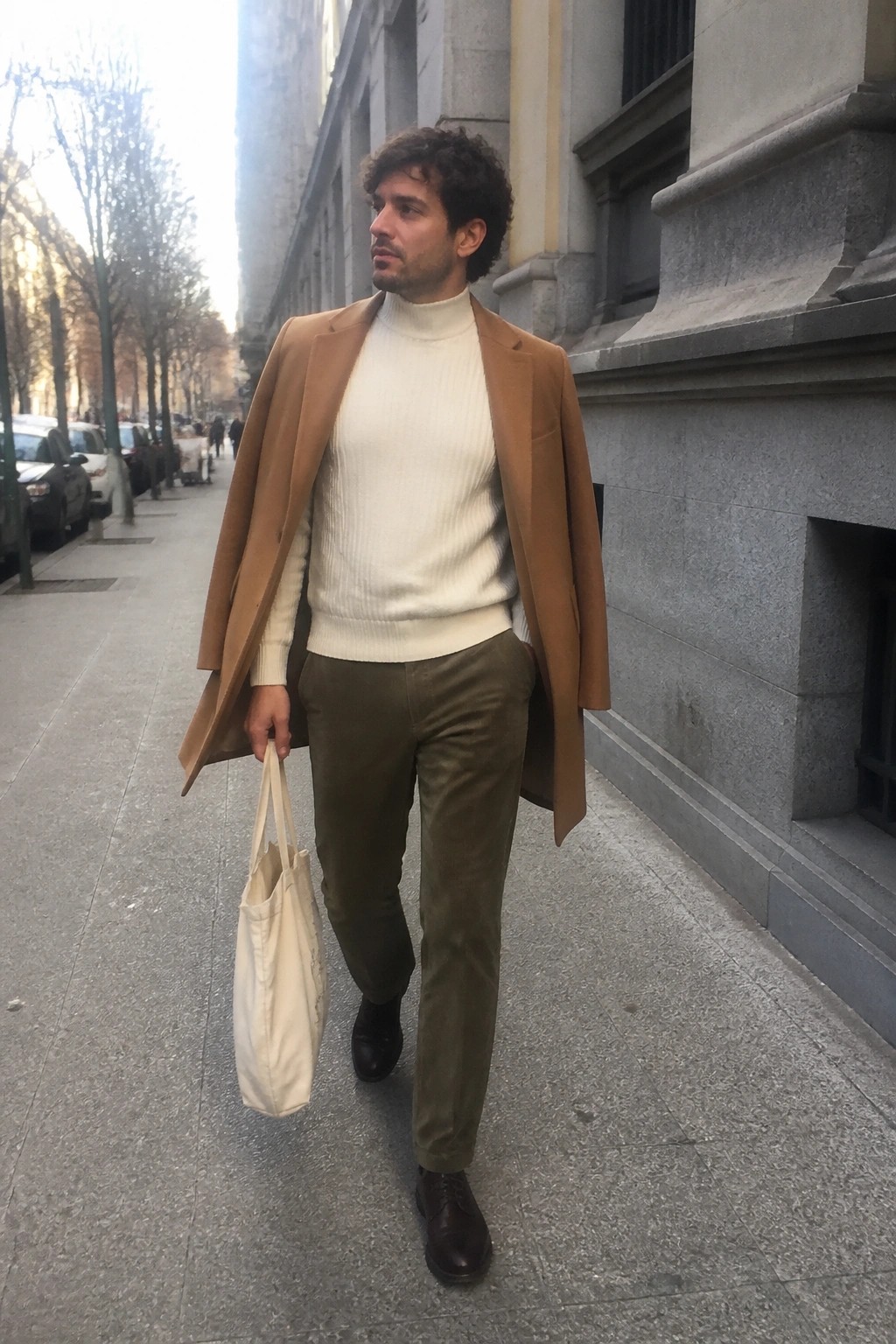 Man in tan overcoat over white turtleneck carrying beige tote on city sidewalk
