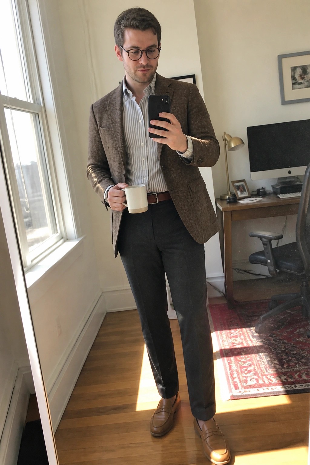 Man in brown textured blazer over striped shirt with gray tailored trousers brown belt and loafers holding a mug and phone in an indoor setting.