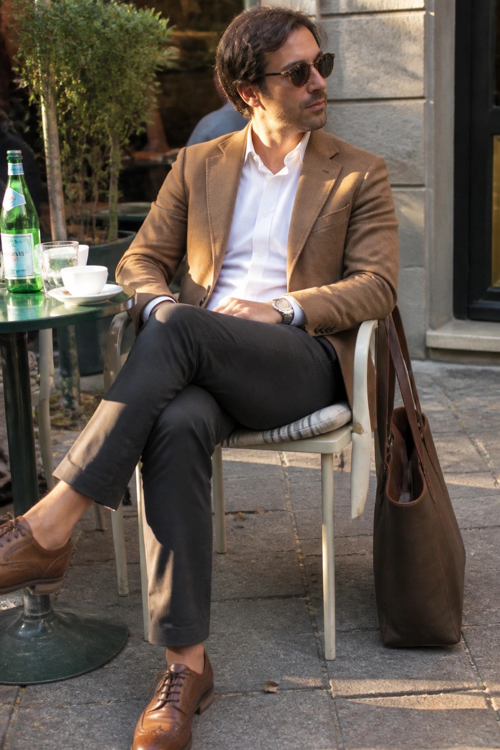 Man in brown blazer and white shirt seated outdoors with leather tote bag