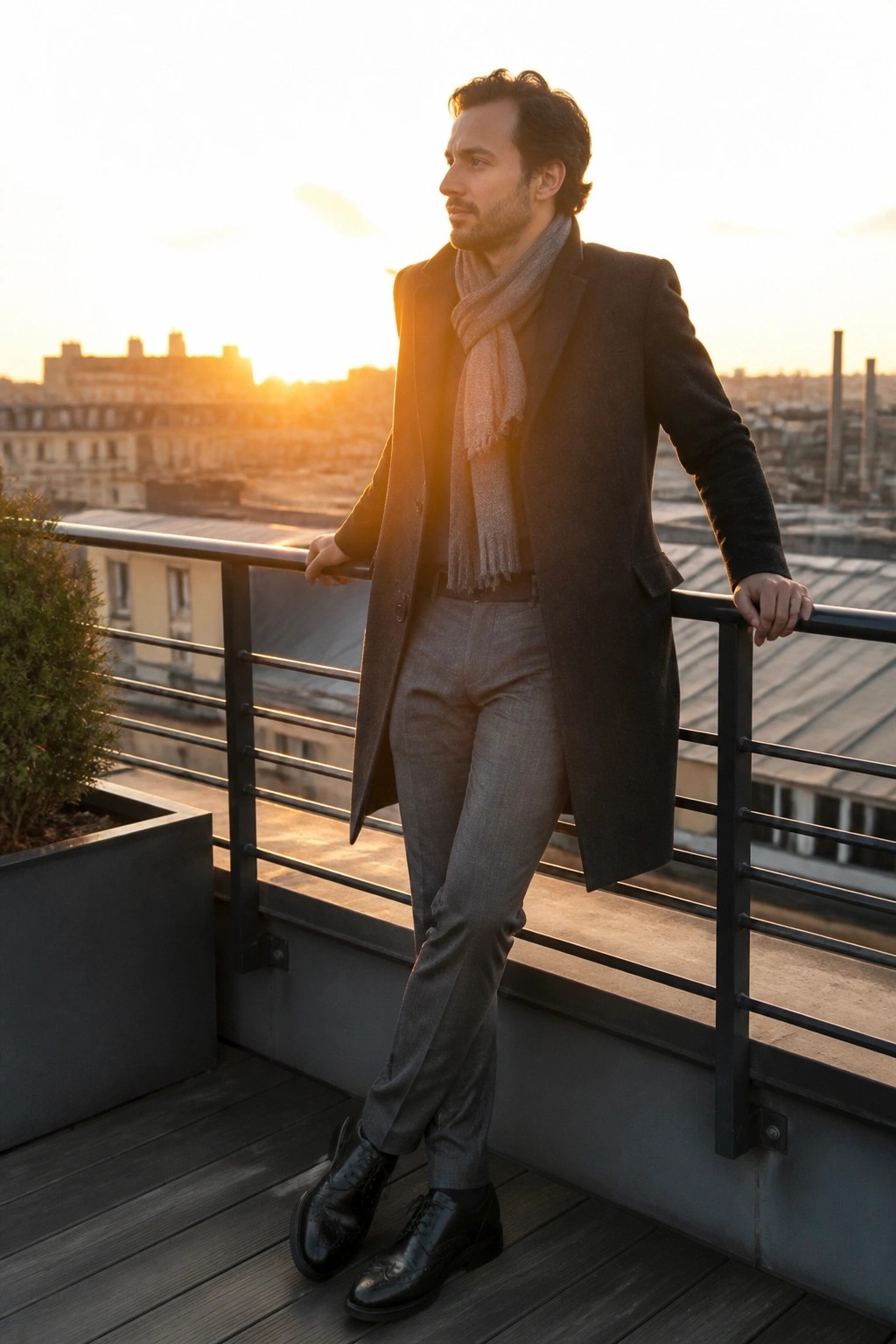 Man in dark overcoat and gray scarf leans on balcony railing at sunset
