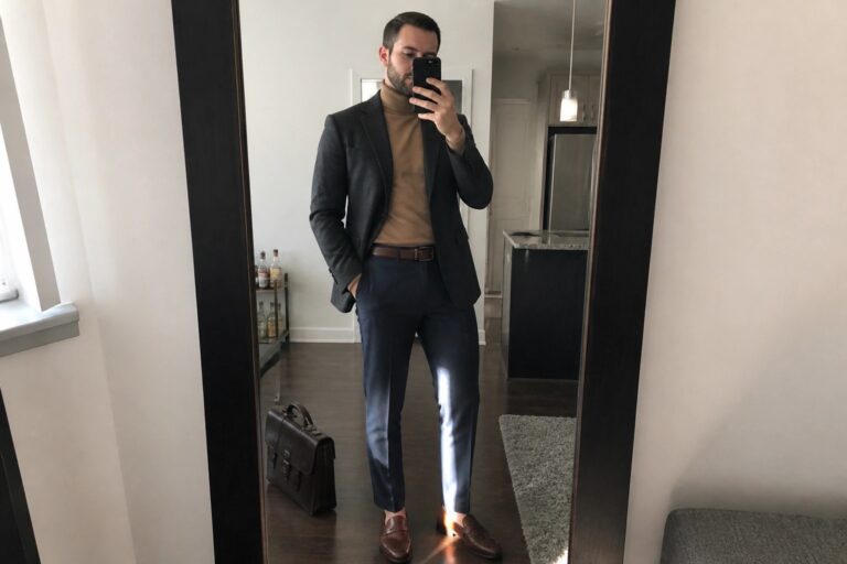 Man in gray blazer and tan turtleneck holding phone and leather bag in mirror