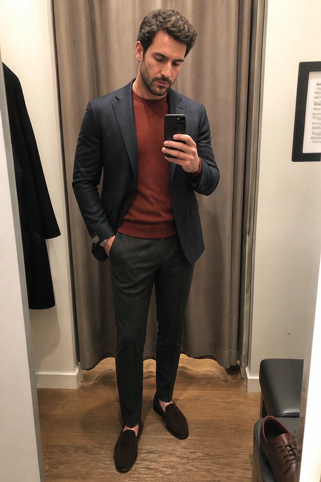 A man wearing a navy blazer over a rust sweater with gray tailored trousers and brown suede loafers taking a mirror selfie in a fitting room.