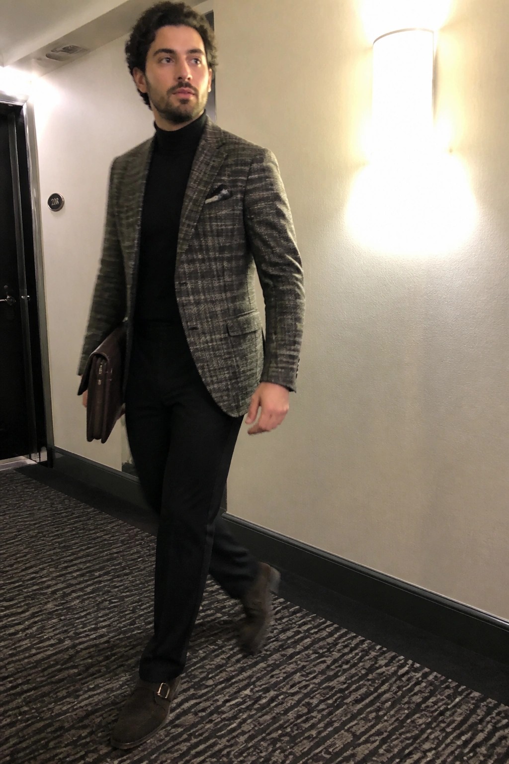 Man in plaid blazer and black turtleneck carrying leather bag in hallway