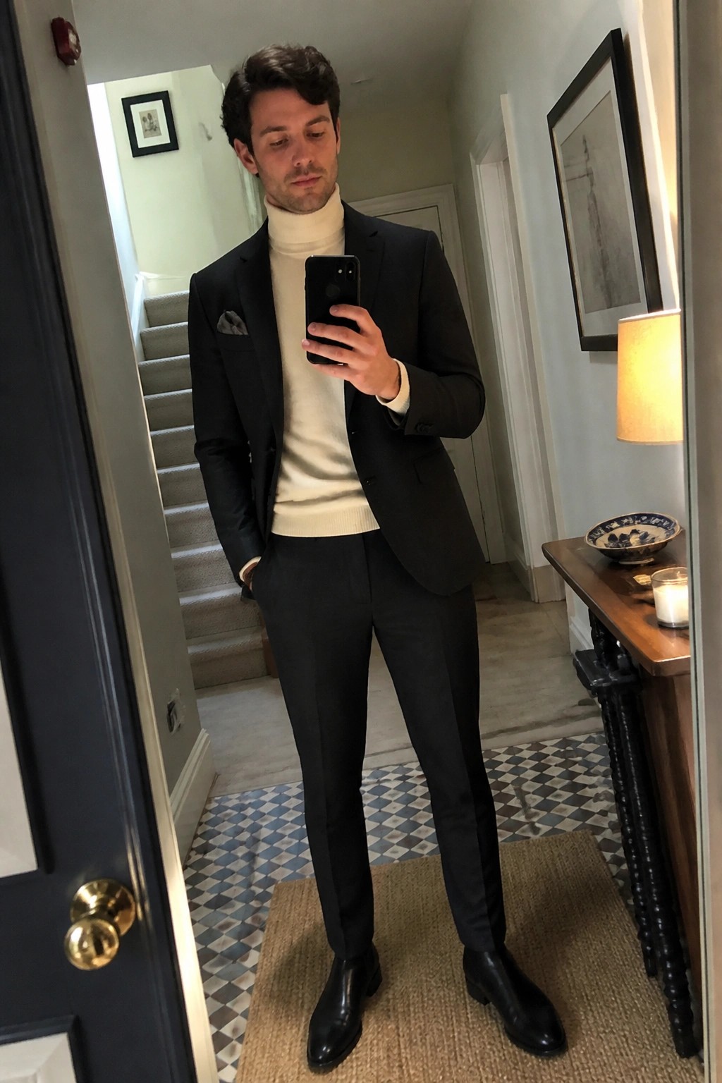 Man in black suit and cream turtleneck takes mirror selfie in tiled hallway