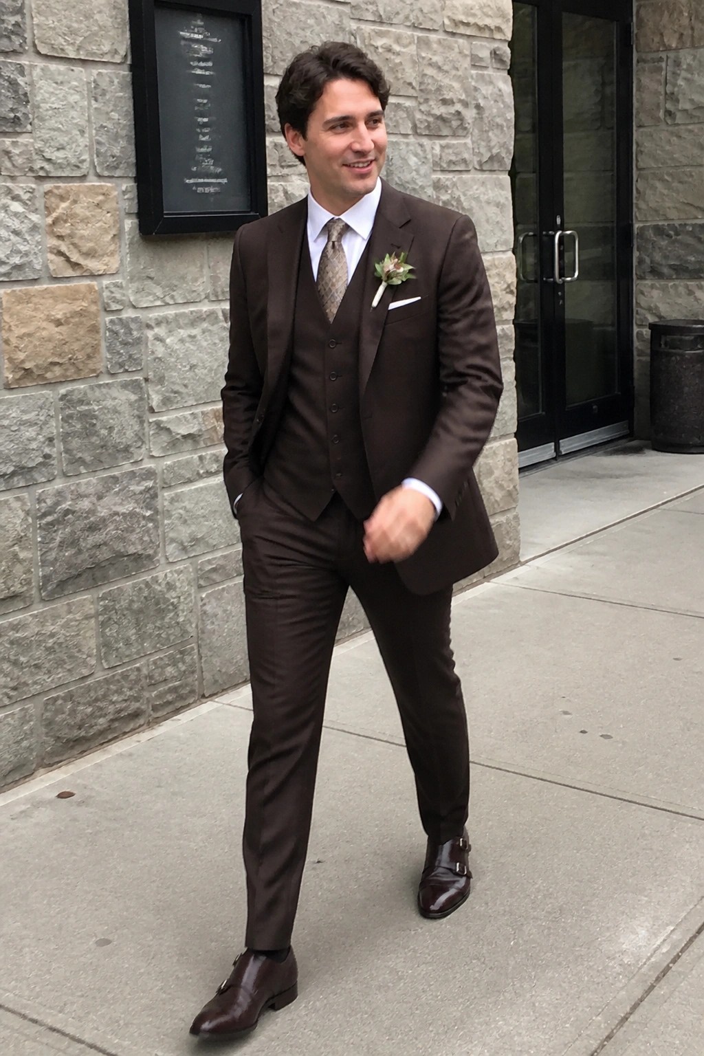 Man in brown three-piece suit with vest, tie, and boutonniere stands outdoors on pavement.