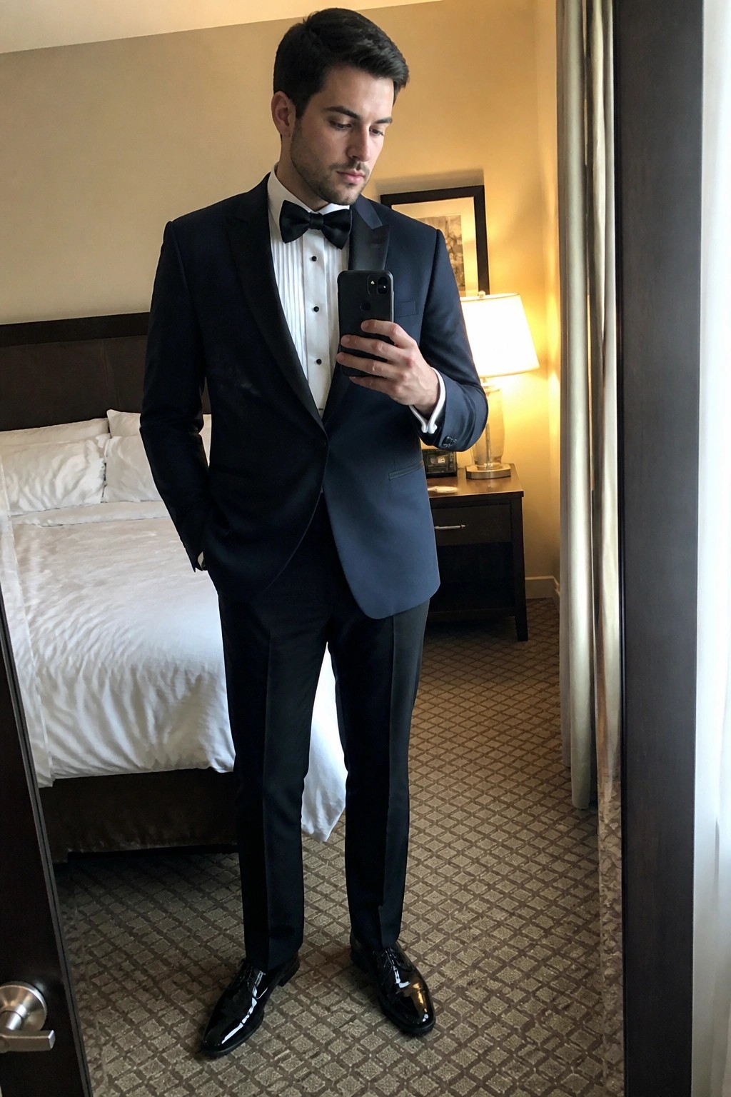Man in black tuxedo and bow tie takes mirror selfie in hotel room.