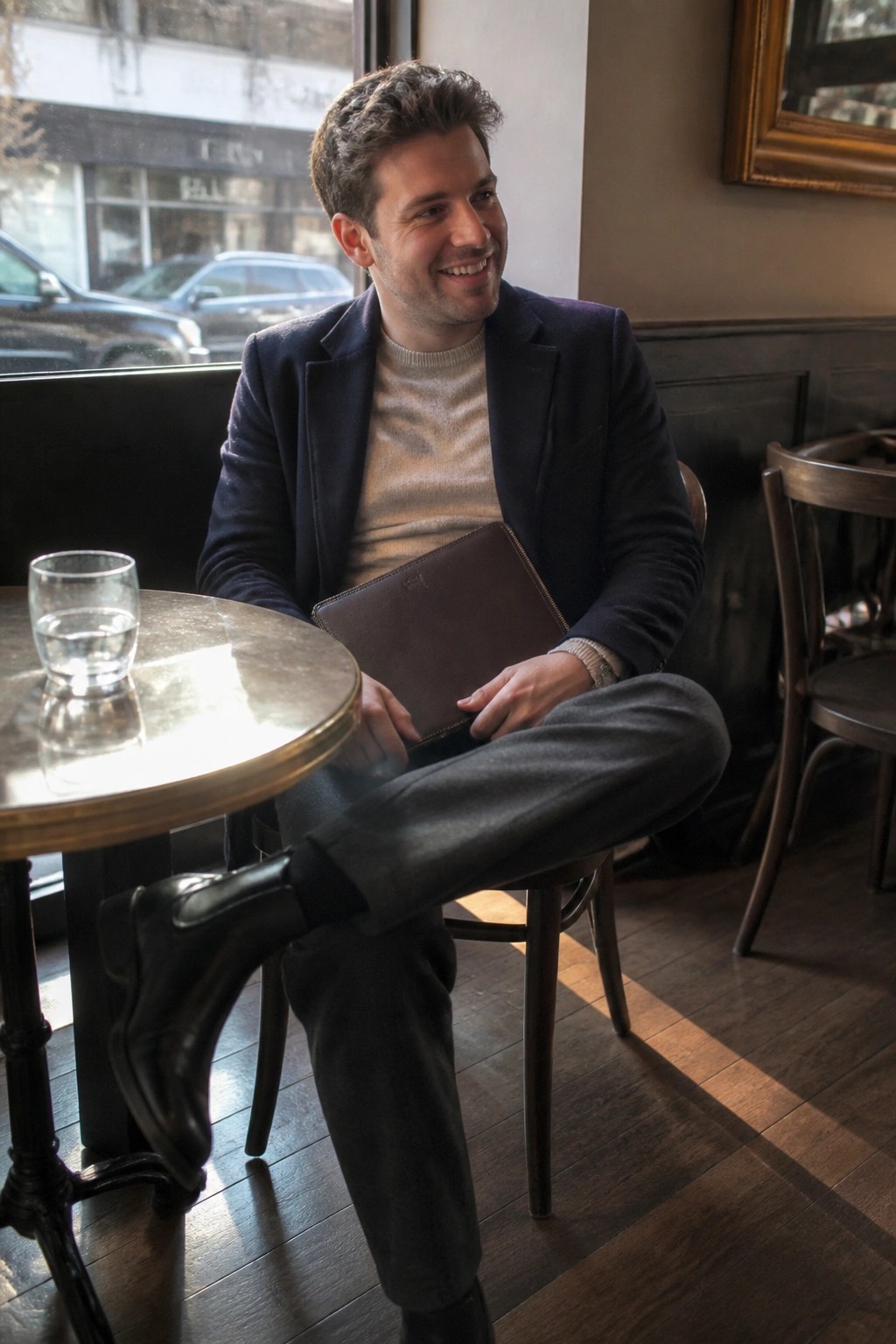 Smiling man in navy blazer holds brown leather pouch at sunny cafe table