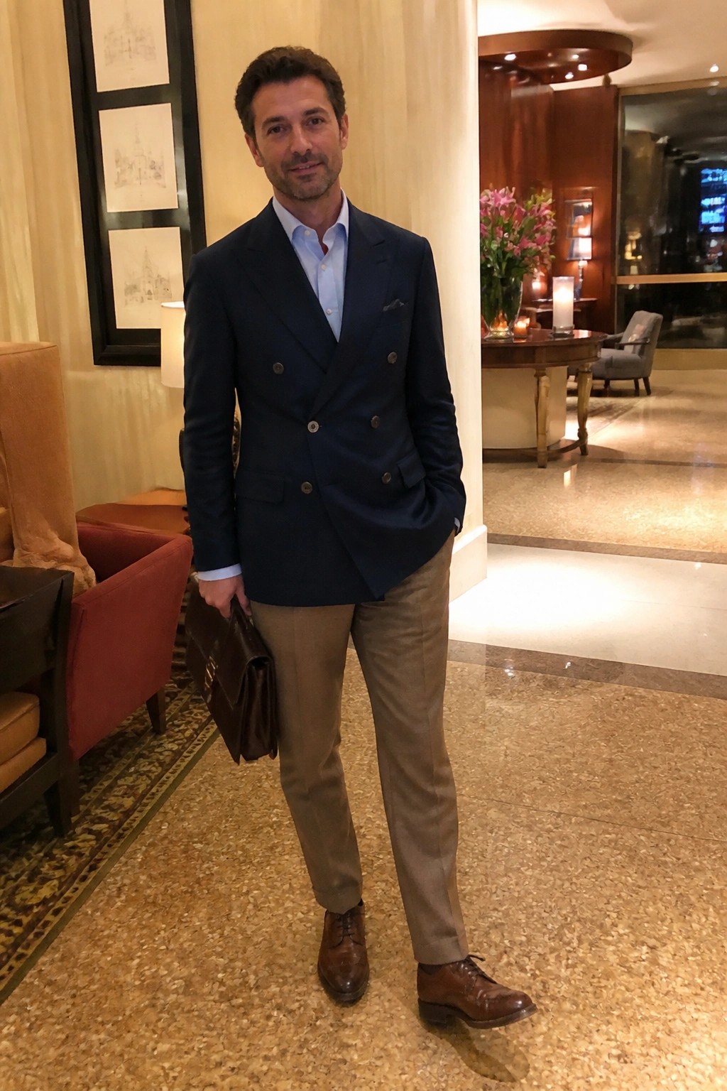 Man in navy double-breasted blazer and khaki pants holding brown briefcase in lobby