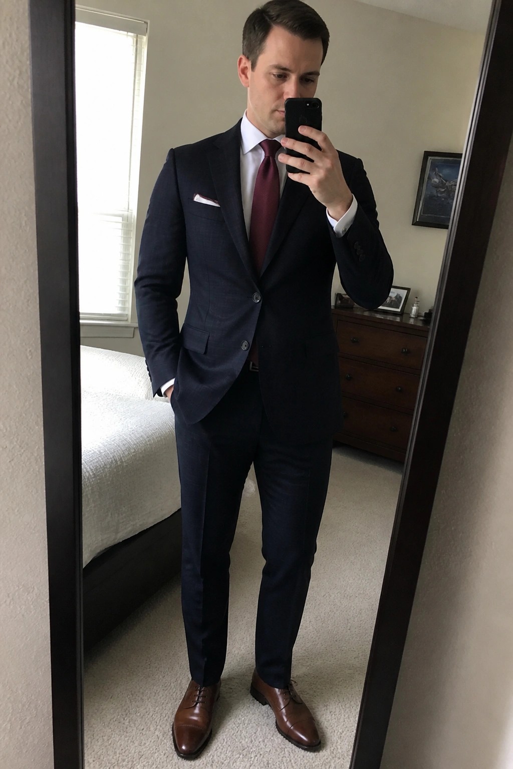Man in navy suit with maroon tie takes bedroom mirror selfie holding phone.