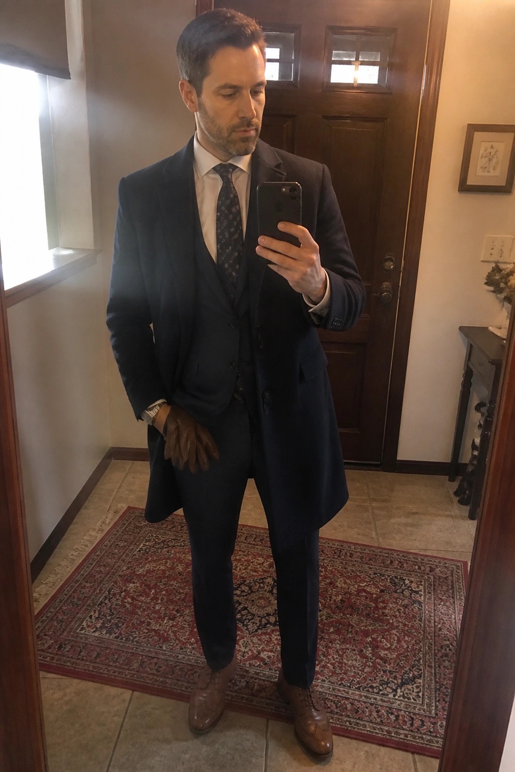 Man in navy three-piece suit taking mirror selfie while holding brown gloves.