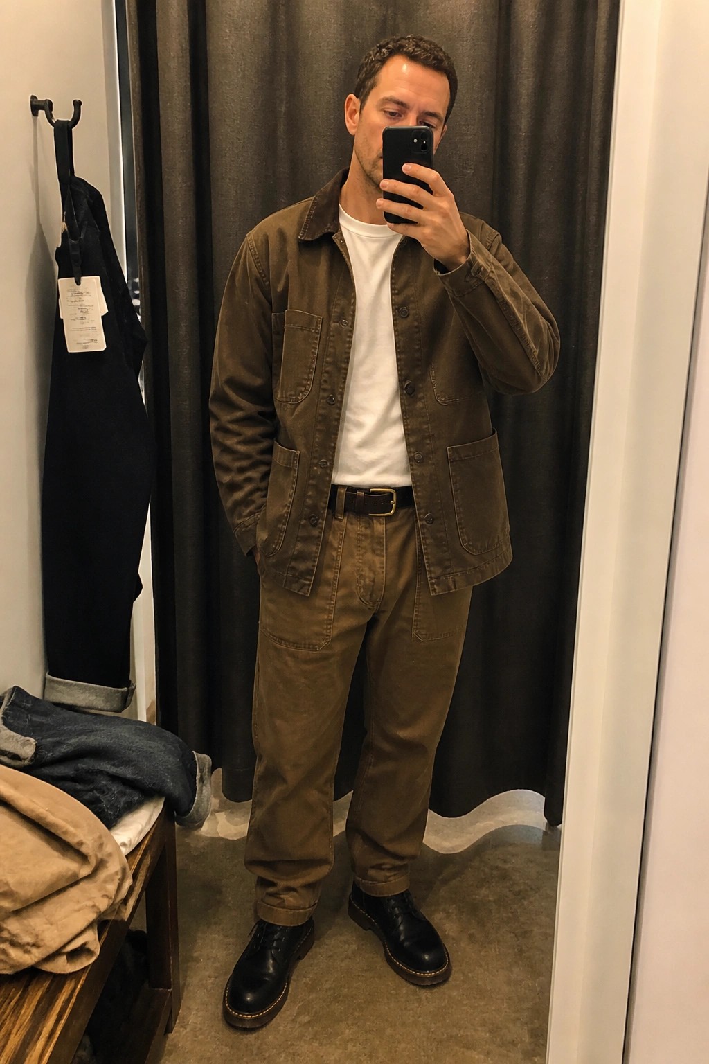 Man in brown utility jacket and pants takes mirror selfie in fitting room.
