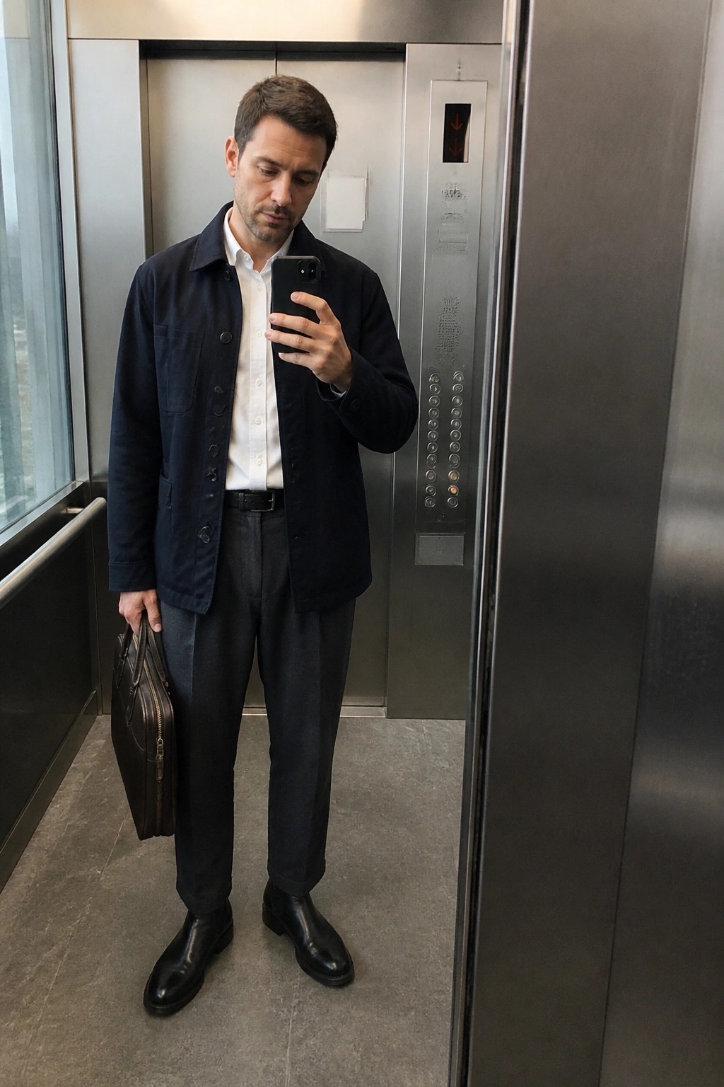 Man in navy jacket and white shirt taking mirror selfie in elevator with briefcase