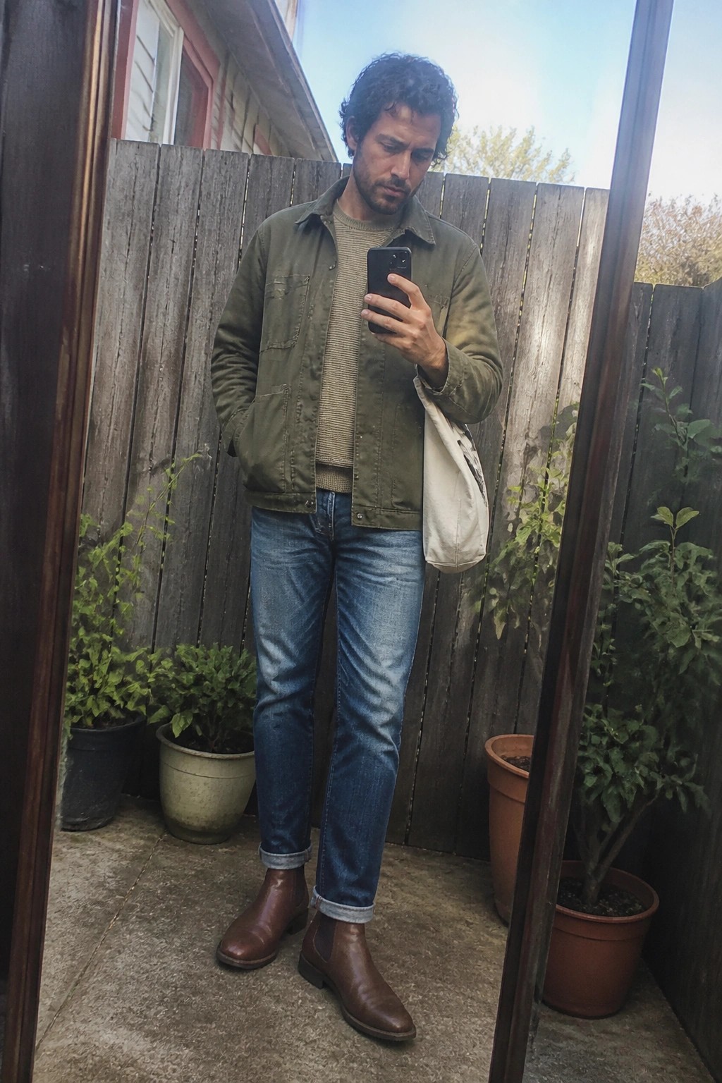 Man in green jacket, striped sweater, blue jeans, and brown boots holding smartphone by fence