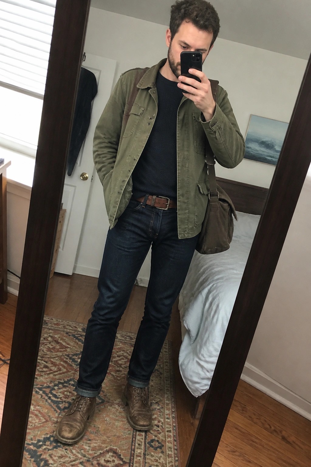 Man in green jacket and jeans taking mirror selfie with brown bag in bedroom