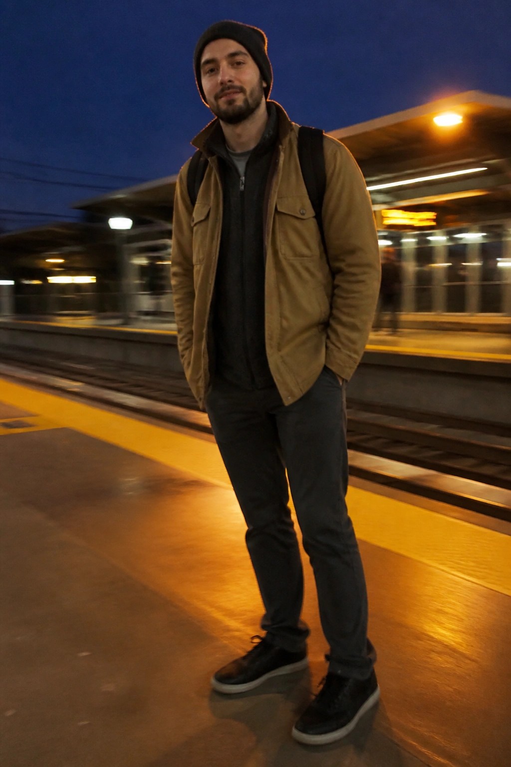 Man in tan jacket and black beanie with backpack on nighttime train platform.