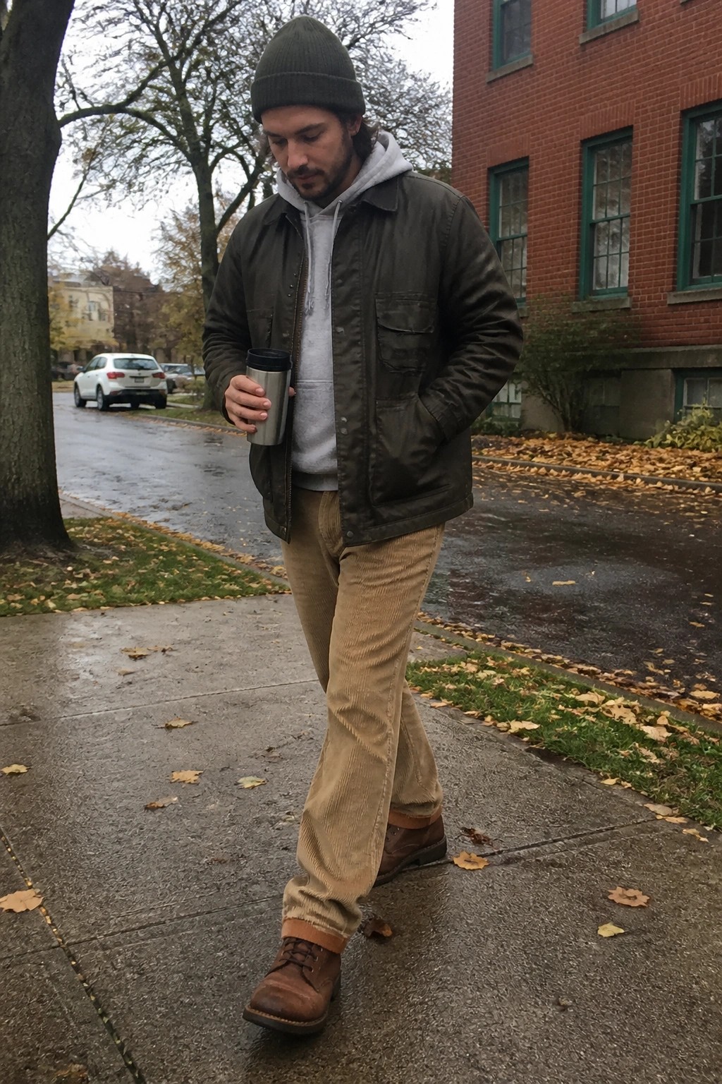 Man in green jacket and beanie walks wet autumn sidewalk holding metal tumbler