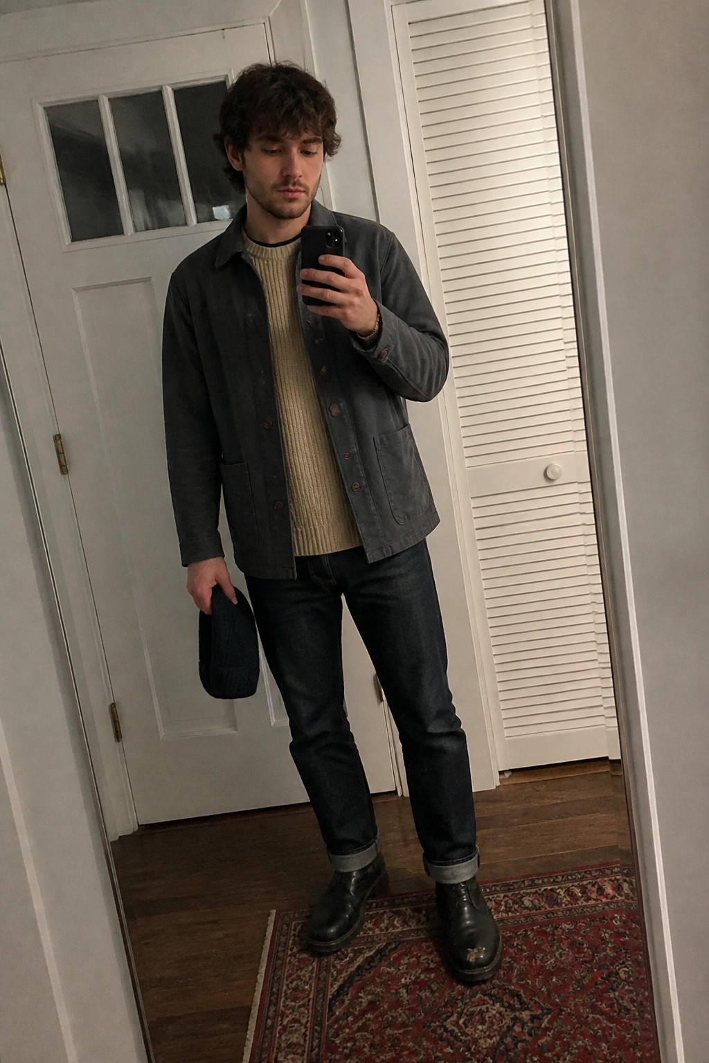 Man in gray jacket over beige sweater taking mirror selfie while holding beanie