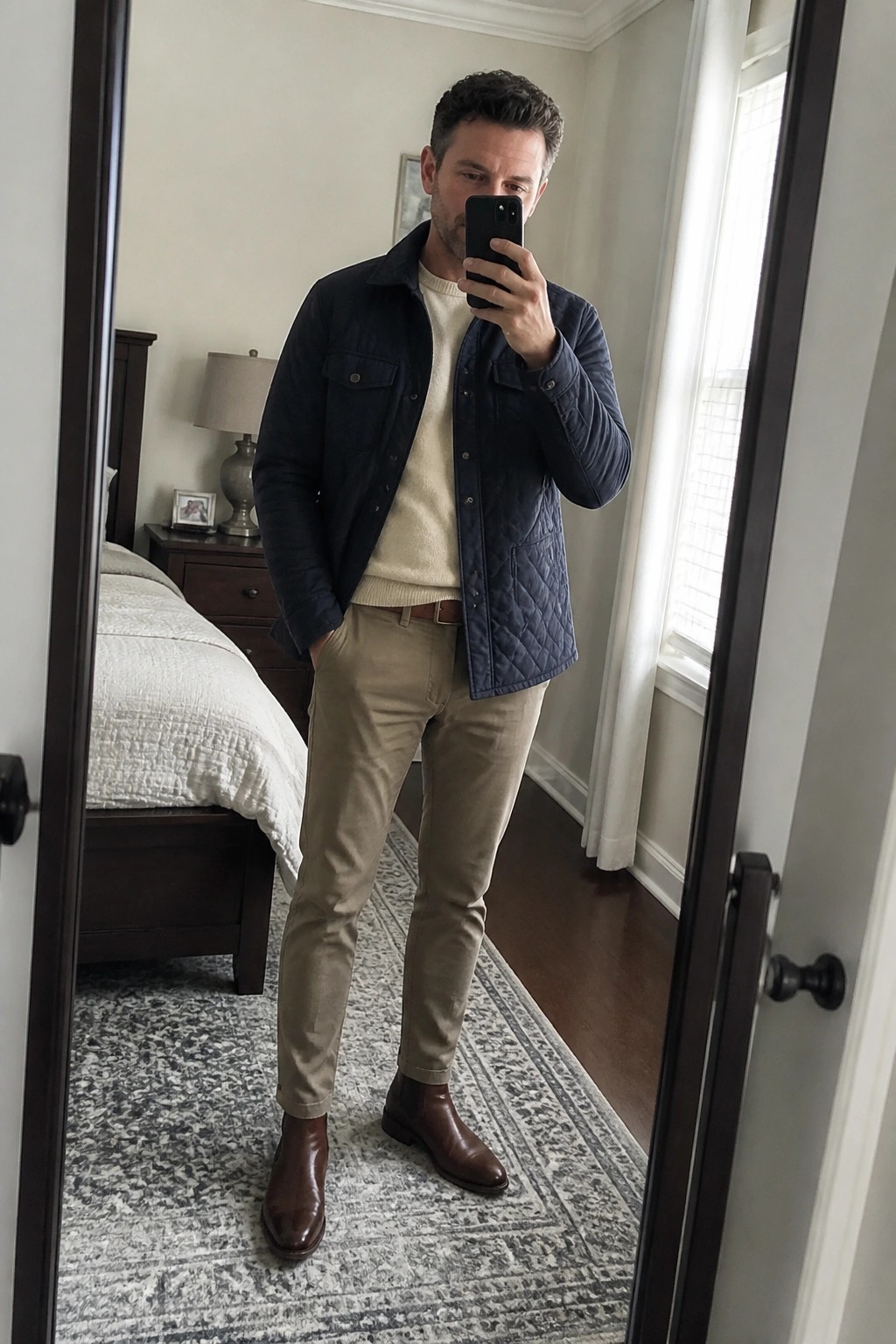 Man in navy quilted jacket, cream sweater, and khaki pants taking mirror selfie in bedroom
