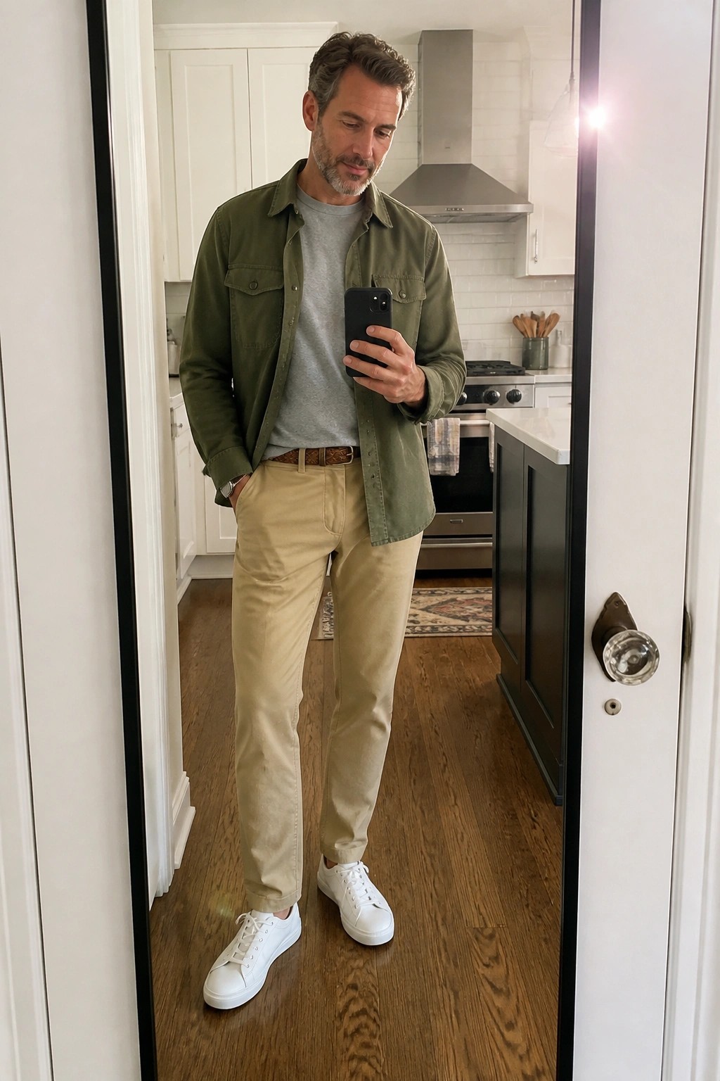 Man in green overshirt and khaki pants taking mirror selfie in kitchen.