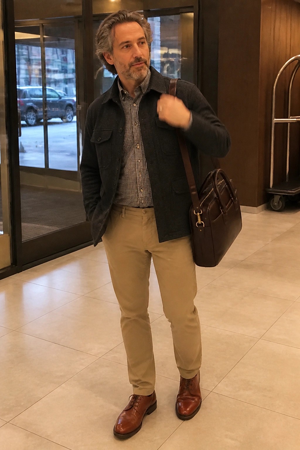 Man wearing dark gray overshirt over gingham shirt tucked into khaki chinos with brown leather boots and matching bag slung over one shoulder.
