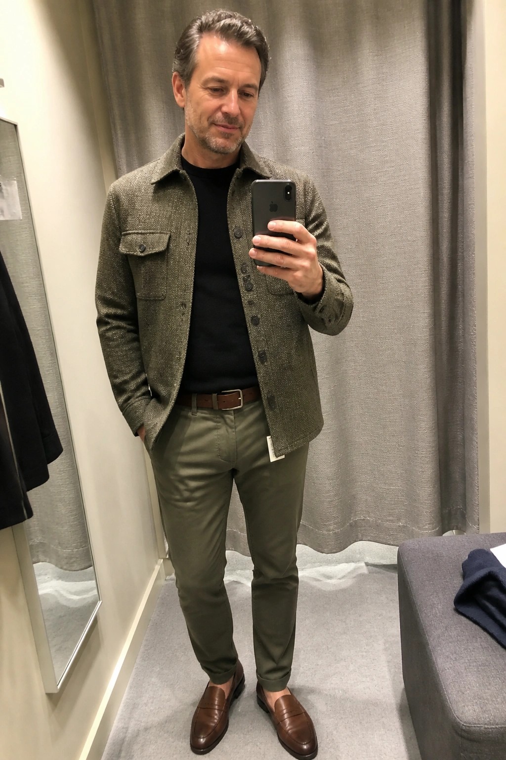 Man in green textured jacket, black shirt, olive pants, brown loafers taking mirror selfie.