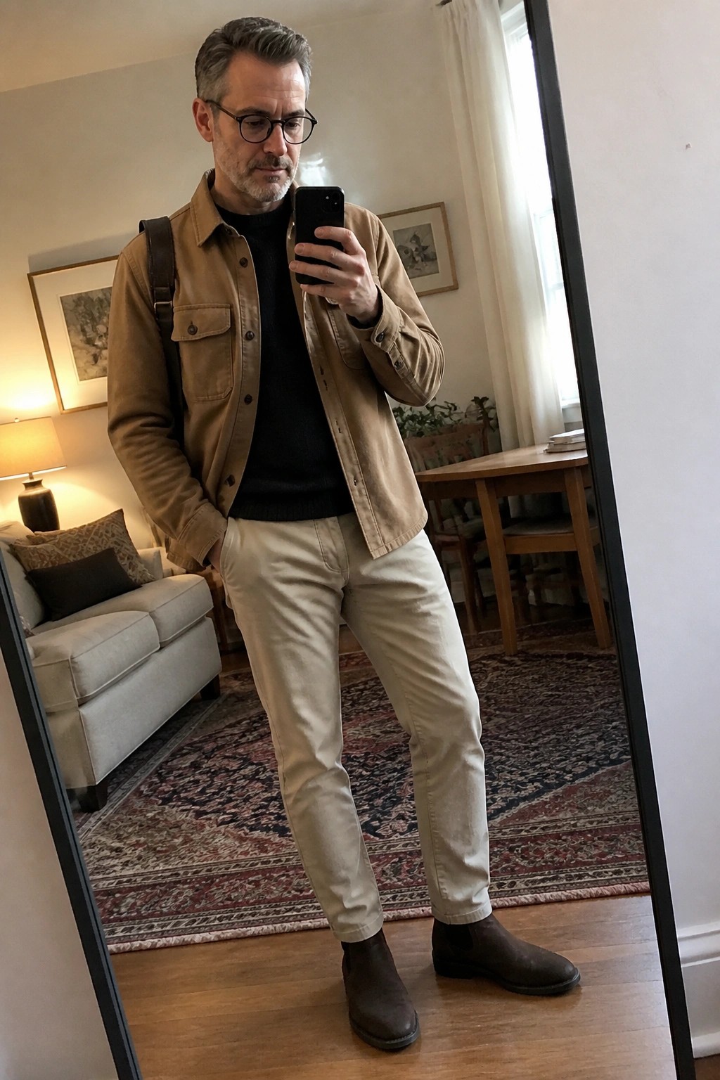 Man in tan jacket and beige pants taking mirror selfie indoors