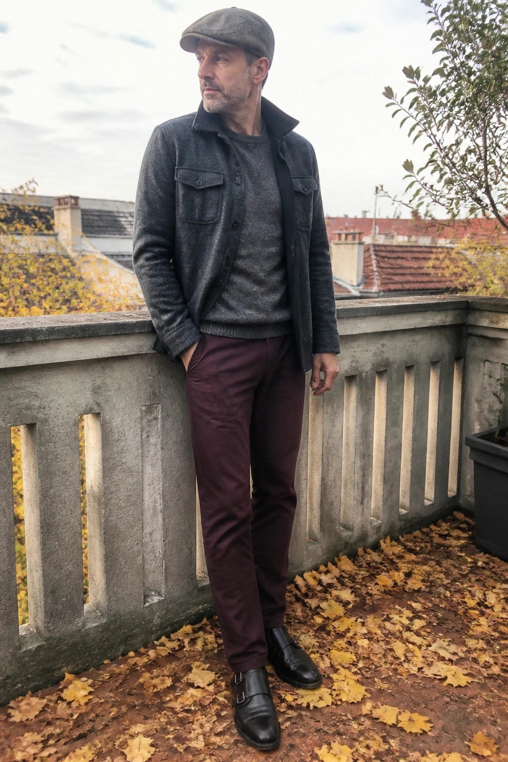 Man in gray cap, jacket, and maroon pants stands on autumn leaf-covered balcony