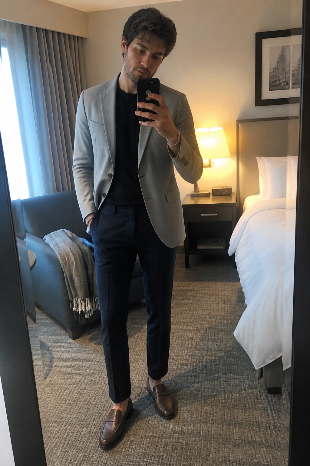 Man in gray blazer and navy pants taking mirror selfie in hotel room