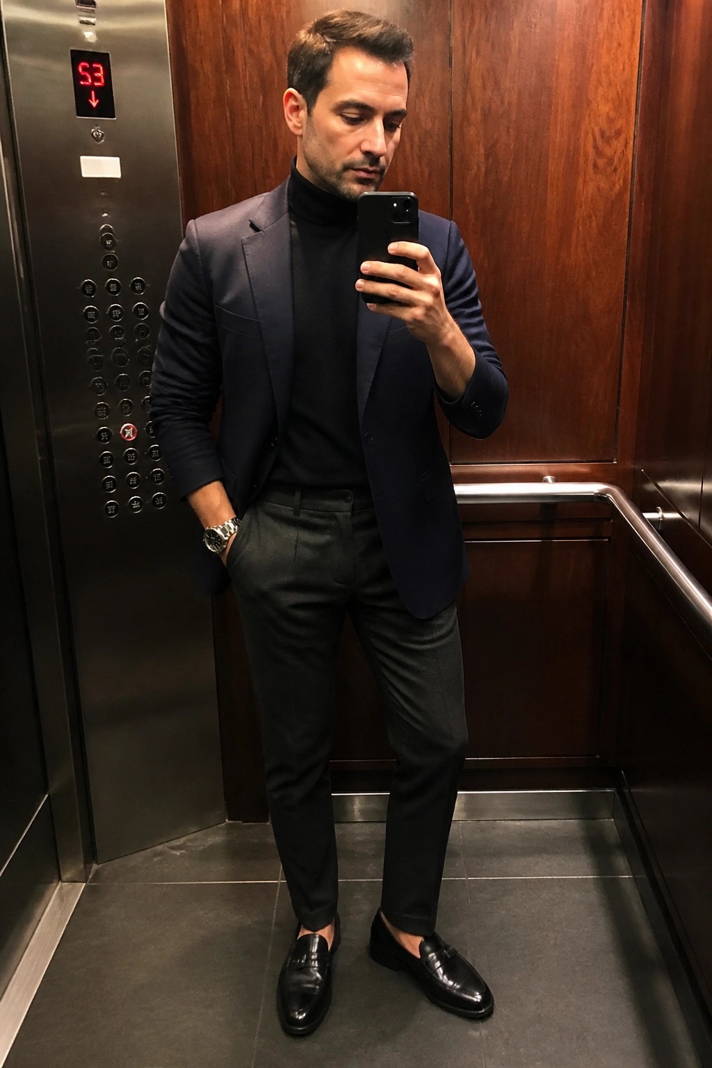 Man in navy blazer and black turtleneck holding phone in elevator