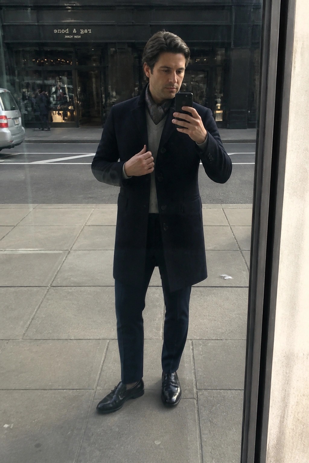 Man in navy overcoat and gray vest views smartphone on city sidewalk.